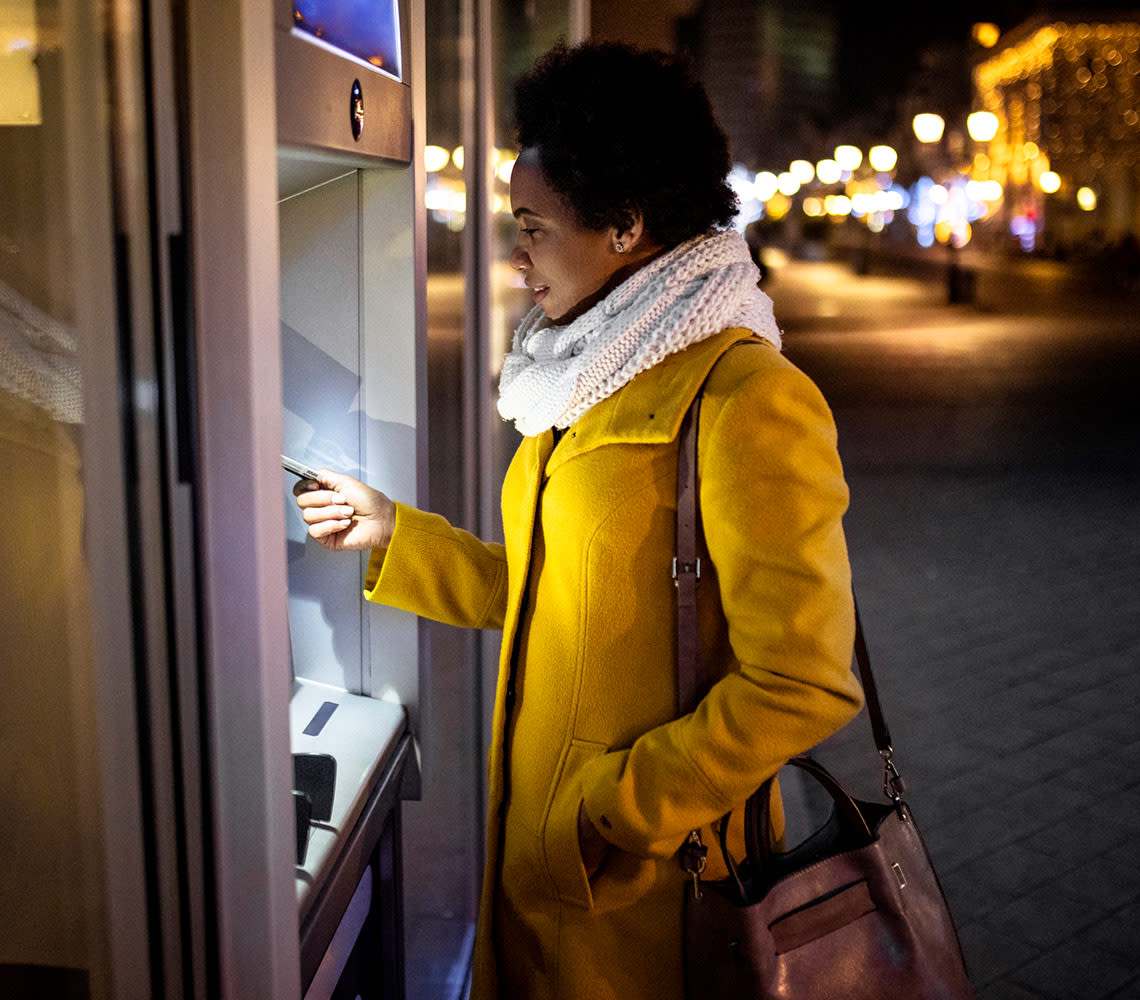 A woman in a yellow coat and white scarf is accessing an ATM, engaged in a transaction in a city landscape.